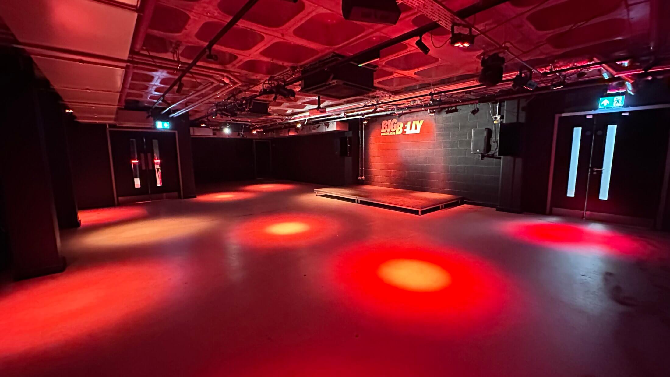 The empty Big Belly performance room, with a black-brick stage, yellow venue logo and red lighting